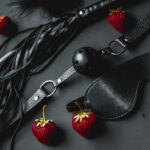 Zdjęcie bdsm. Czarna maska, flogger (bicz), knebel i truskawski.
