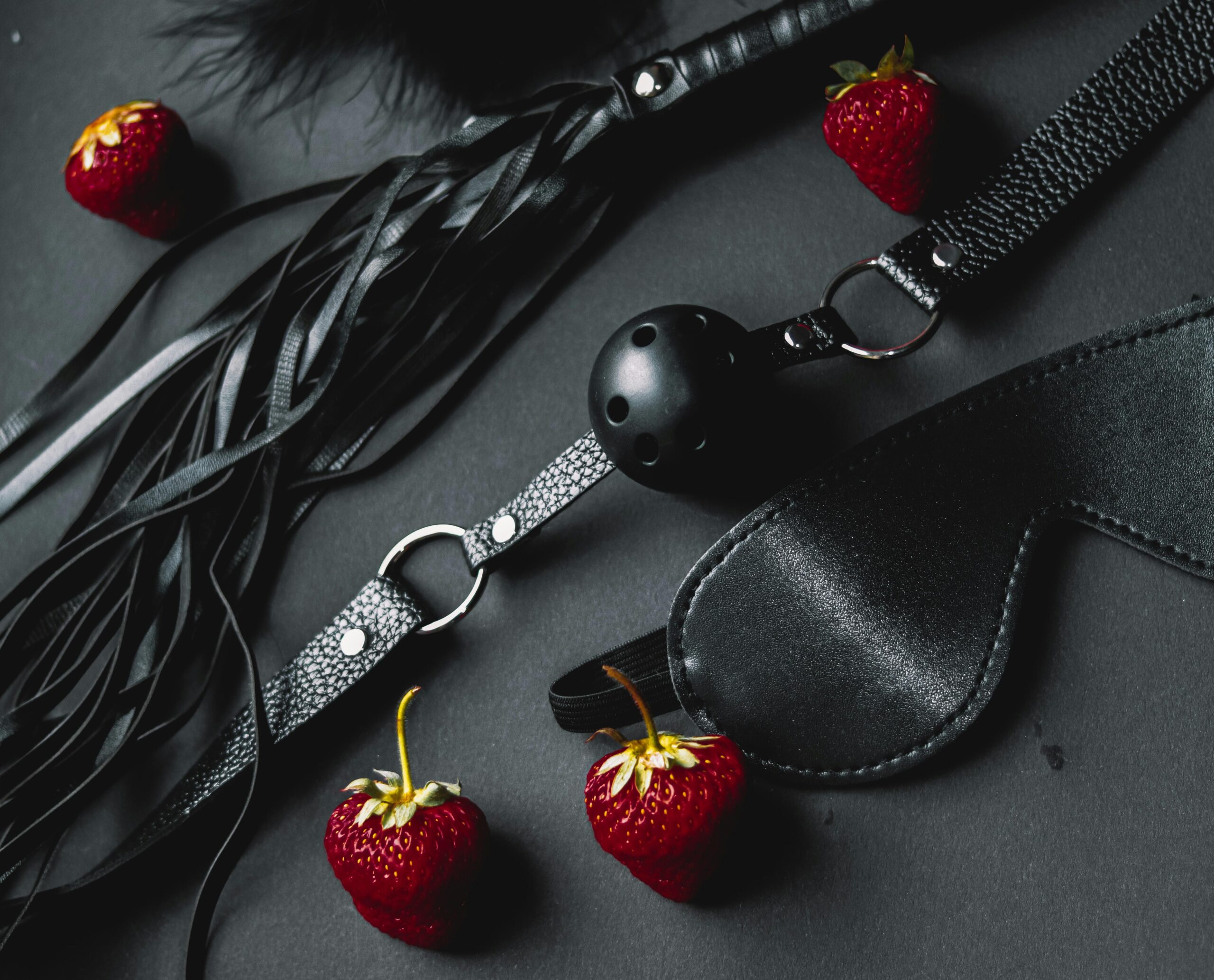 Zdjęcie bdsm. Czarna maska, flogger (bicz), knebel i truskawski.